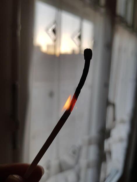 A person's hand holding an incense stick near a window frame, with the smoke showing a clear draft in a dimly lit room, illustrating a DIY draft test.